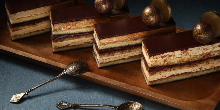 4 very neatly sliced chocolate and custard slices decorated with gold-dusted chocolate balls and a coin sitting on a wooden platter with antique silver dessert spoons.